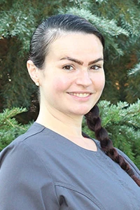 Woman in gray scrubs smiling outdoors with green trees in the background.