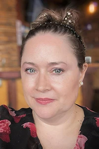 Woman with light eyes and floral top smiling indoors with blurred background.